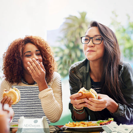 Two women eating burgers, wearing NEATsheets and using NEATwipes at a tailgate.