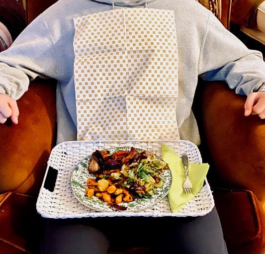 Man sitting in a chair eating dinner in front of a TV wearing a Khaki Gingham NEATsheet, the ideal disposable clothing protector..