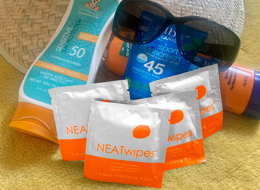NEATwipes Fresh Citrus handwipes on a beach towel with sunscreen, a hat, and sunglasses.